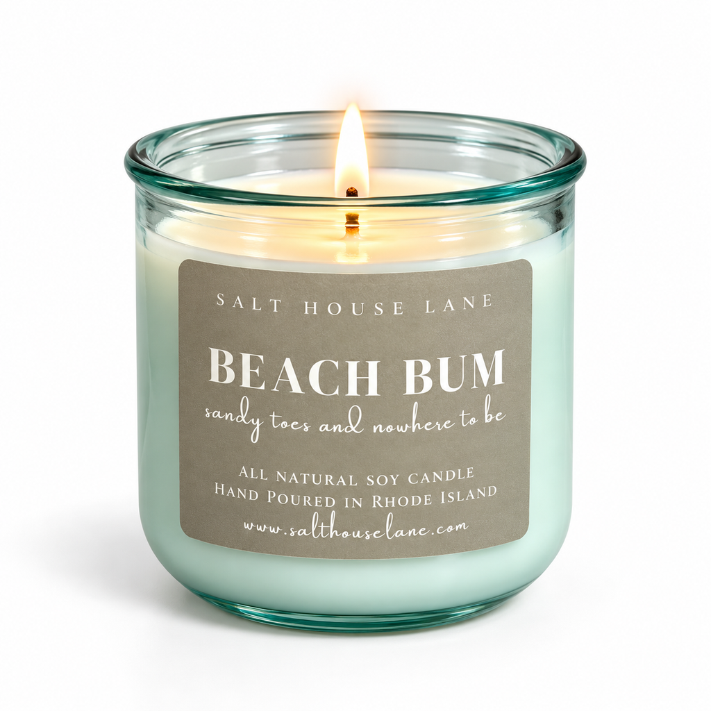 A lit Salt House Lane 'Beach Bum' soy candle in a clear glass jar with a mint green wax and a brown paper label, set against a plain white background.