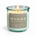 A lit Salt House Lane 'Beach Bum' soy candle in a clear glass jar with a mint green wax and a brown paper label, set against a plain white background.