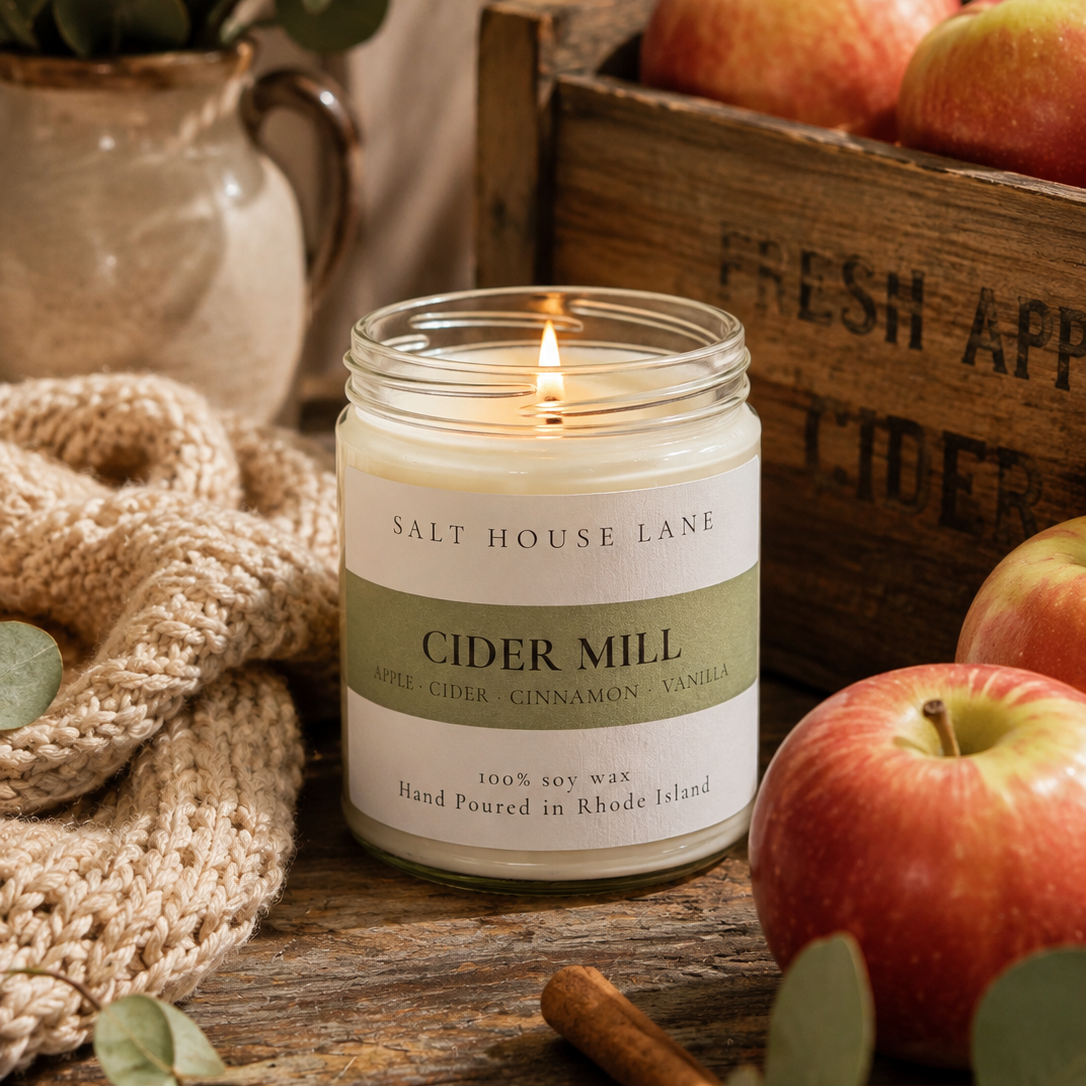 A lit jar candle labeled 'CIDER MILL' by Salt House Lane sits on a rustic wooden table surrounded by red apples, eucalyptus leaves, cinnamon sticks, and a knitted blanket.
