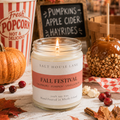 A lit soy candle in a clear glass jar labeled 'SALT HOUSE LANE' and 'FALL FESTIVAL' sits on a rustic white wooden table surrounded by autumn props including a pumpkin, popcorn bucket, candy apple, cranberries, and fall leaves.