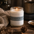 A lit candle in a clear glass jar with a cream label reading 'SALT HOUSE LANE' and 'MIDNIGHT MOON,' styled on a rustic wooden surface with pinecones, dried leaves, a knit blanket, and a book in the background.