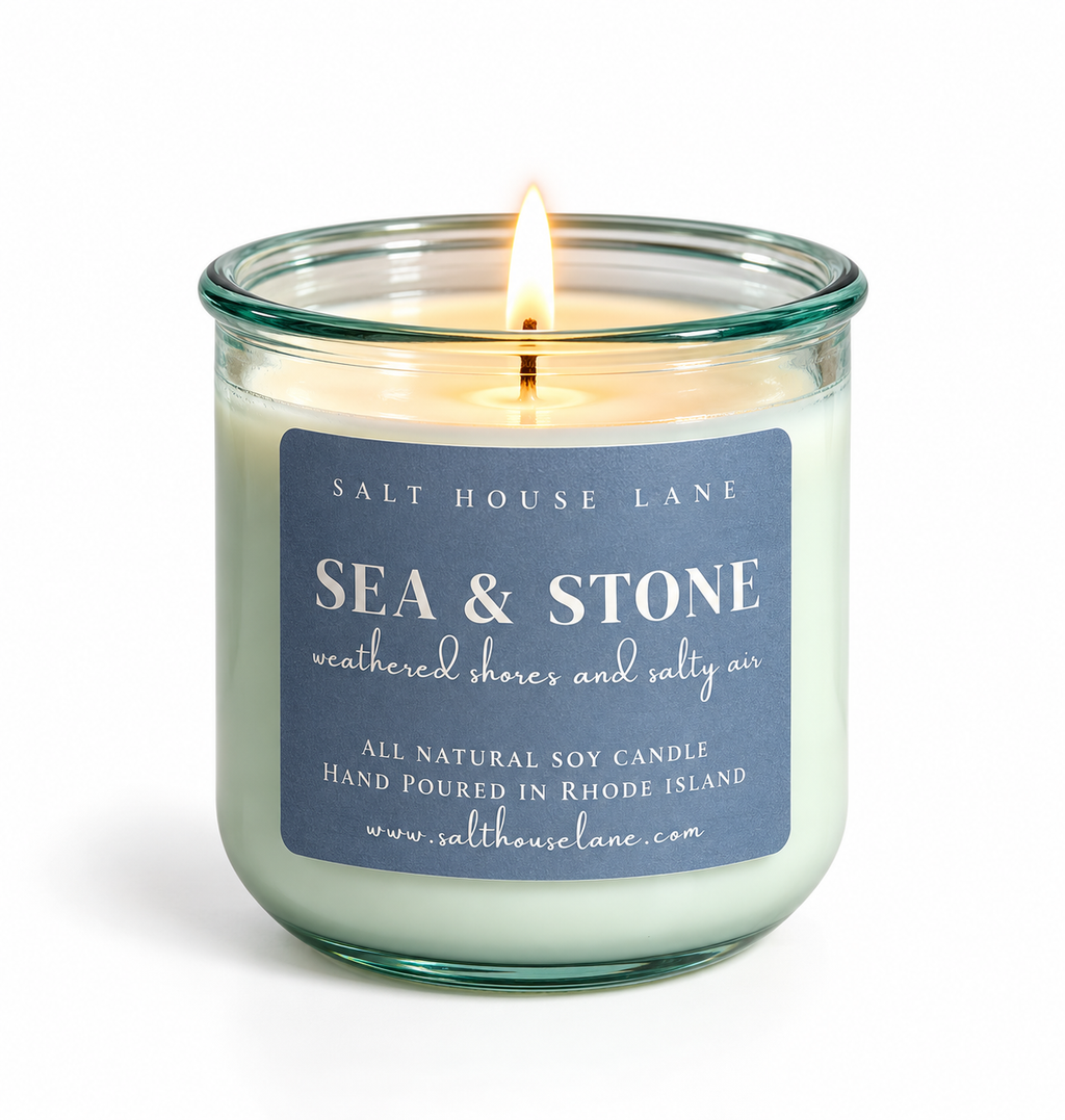 A lit salt house candle in a clear glass jar with a blue label reading 'SEA & STONE' and 'weathered shores and salty air' by Salt House Lane, shown on a white background.