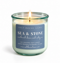 A lit salt house candle in a clear glass jar with a blue label reading 'SEA & STONE' and 'weathered shores and salty air' by Salt House Lane, shown on a white background.