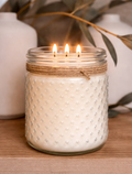 White textured candle in a glass jar with twine, lit on a wooden surface with decorative elements.