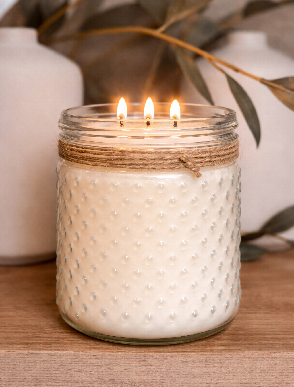 White textured candle in a glass jar with twine, lit on a wooden surface with decorative elements.