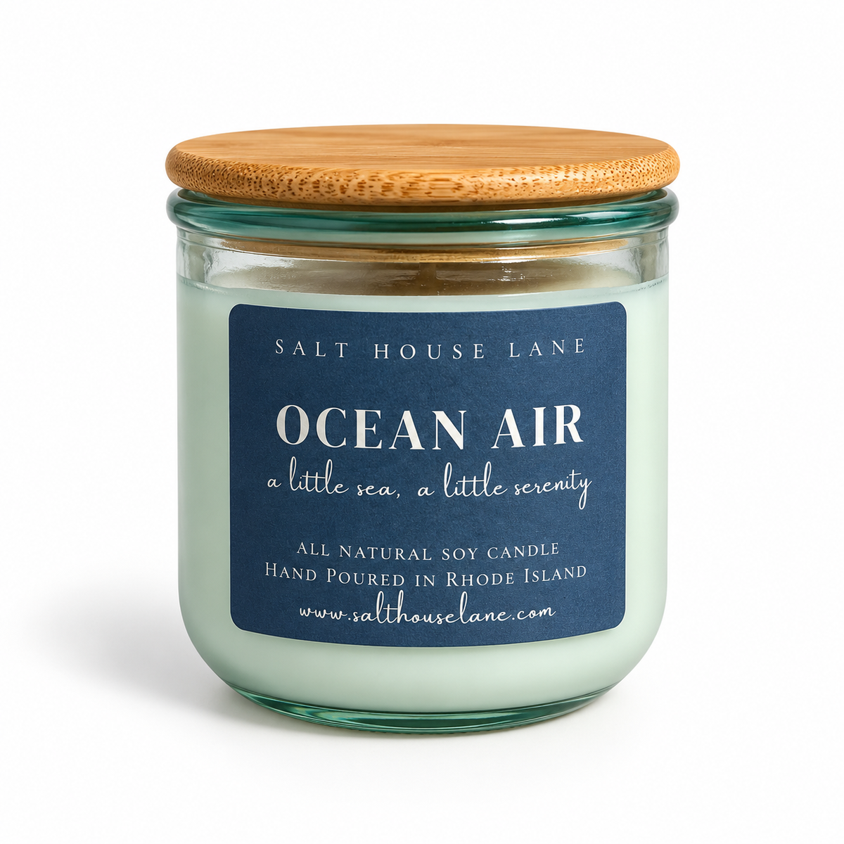 A glass jar candle with pale mint wax, a light wood lid, and a dark blue label reading 'OCEAN AIR' by Salt House Lane, shown on a white background.