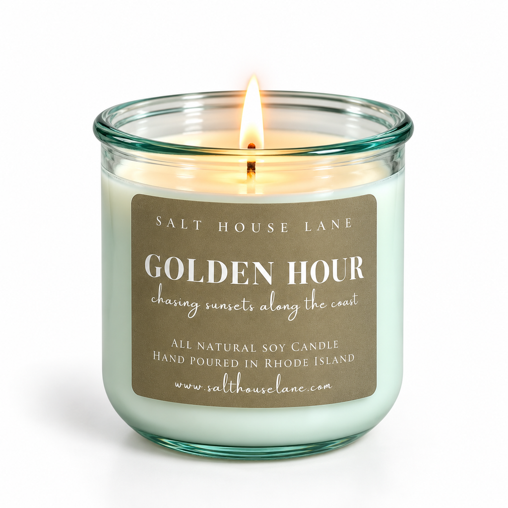 A lit soy candle in a clear glass jar with a teal-green tint, featuring a brown paper label that reads 'GOLDEN HOUR' by Salt House Lane. The candle is set against a clean white background.