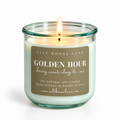 A lit soy candle in a clear glass jar with a teal-green tint, featuring a brown paper label that reads 'GOLDEN HOUR' by Salt House Lane. The candle is set against a clean white background.
