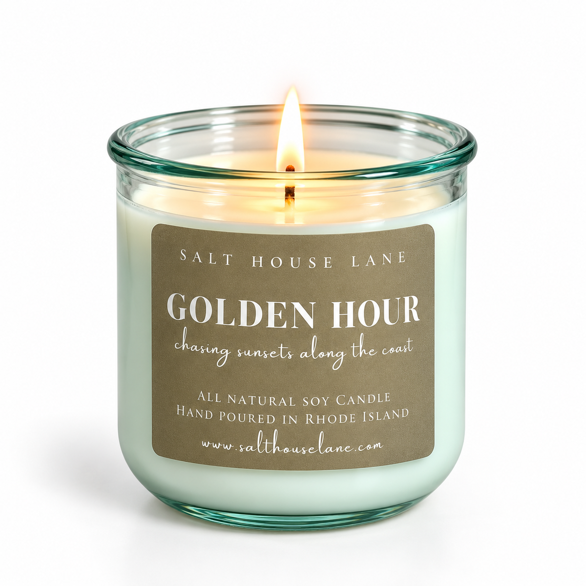 A lit soy candle in a clear glass jar with a teal-green tint, featuring a brown paper label that reads 'GOLDEN HOUR' by Salt House Lane. The candle is set against a clean white background.